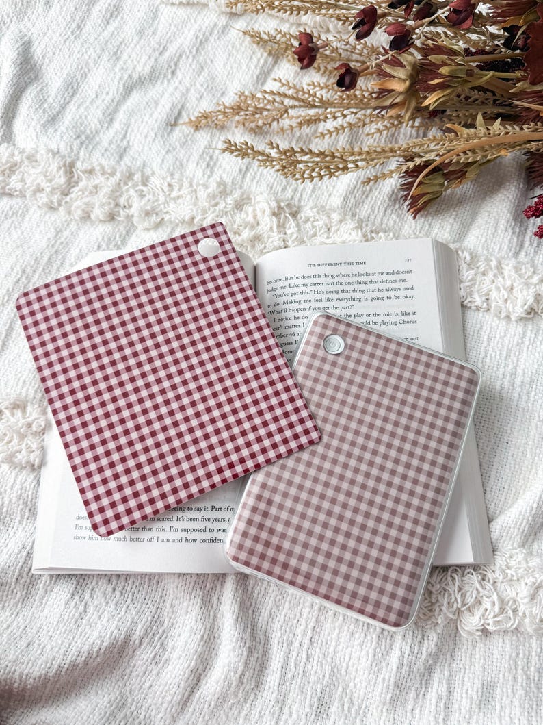May include: Two square coasters featuring a red and white gingham check pattern. Each coaster has a small white circular accent in the corner. The coasters are positioned on top of an open book with visible printed text on the pages.