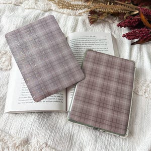 May include: Two rectangular e-reader cases featuring a muted mauve and grey plaid pattern. One case is detached, showing a subtle holographic star overlay, while the other is a clear-framed case fitted onto a device resting on an open book.
