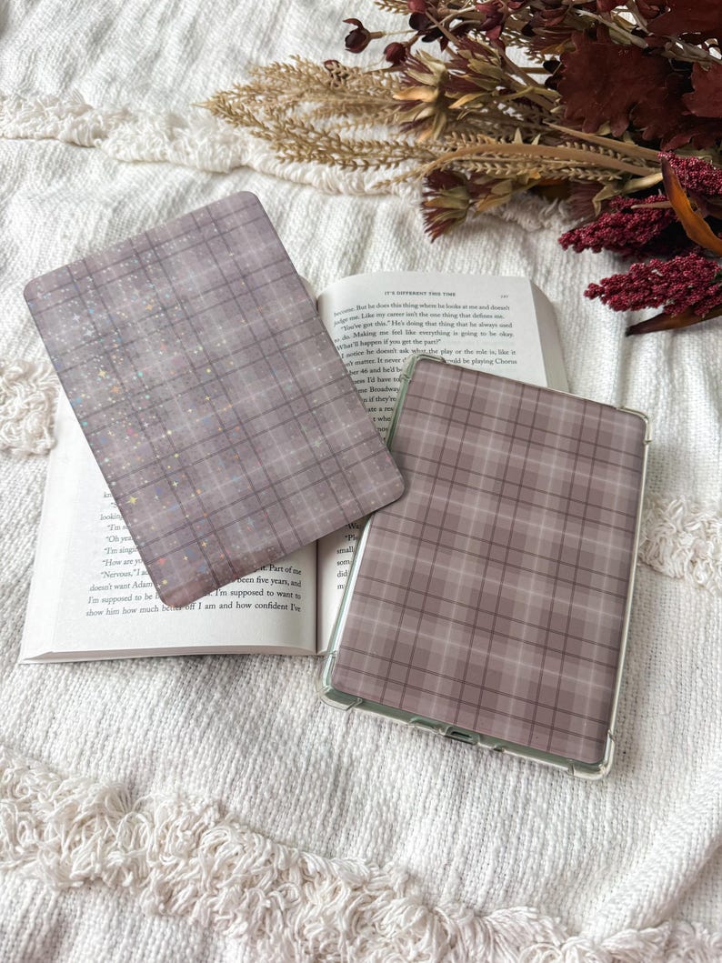 May include: Two rectangular e-reader cases featuring a muted mauve and grey plaid pattern. One case is detached, showing a subtle holographic star overlay, while the other is a clear-framed case fitted onto a device resting on an open book.