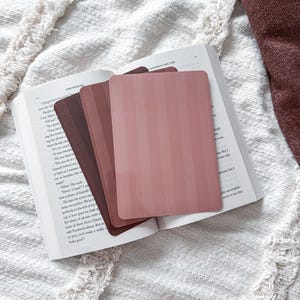 May include: A set of three rectangular bookmarks with rounded corners, featuring a vertical striped pattern in varying shades of dusty rose, mauve, and deep burgundy, resting on the open pages of a printed book with visible black text.