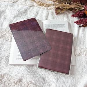 May include: Two rectangular e-reader or tablet cases. One case features a dark purple surface with a fine, multi-colored glitter finish. The second case is encased in a clear protective shell and displays a dark maroon and black plaid pattern.