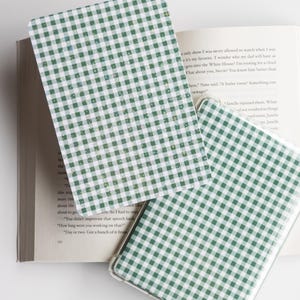 May include: Green and white gingham pattern on a phone case and a bookmark. The phone case has a clear border. The bookmark and phone case are resting on an open book with text.