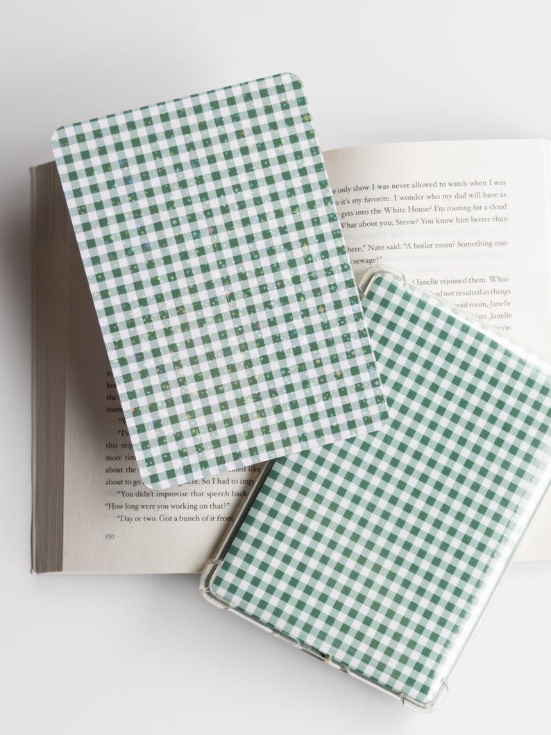 May include: Green and white gingham pattern on a phone case and a bookmark. The phone case has a clear border. The bookmark and phone case are resting on an open book with text.