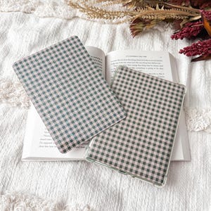 May include: Two rectangular e-reader cases featuring a black and light pink gingham check pattern. One case is positioned flat, while the other is slightly angled, both displaying a consistent grid design across their surfaces.