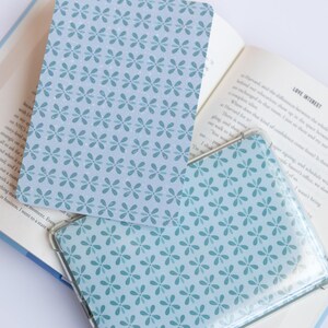 May include: Two light blue floral patterned cards, one inside a clear plastic sleeve, rest on an open book. The cards and sleeve feature a repeating pattern of small, stylized flowers in a darker shade of blue.