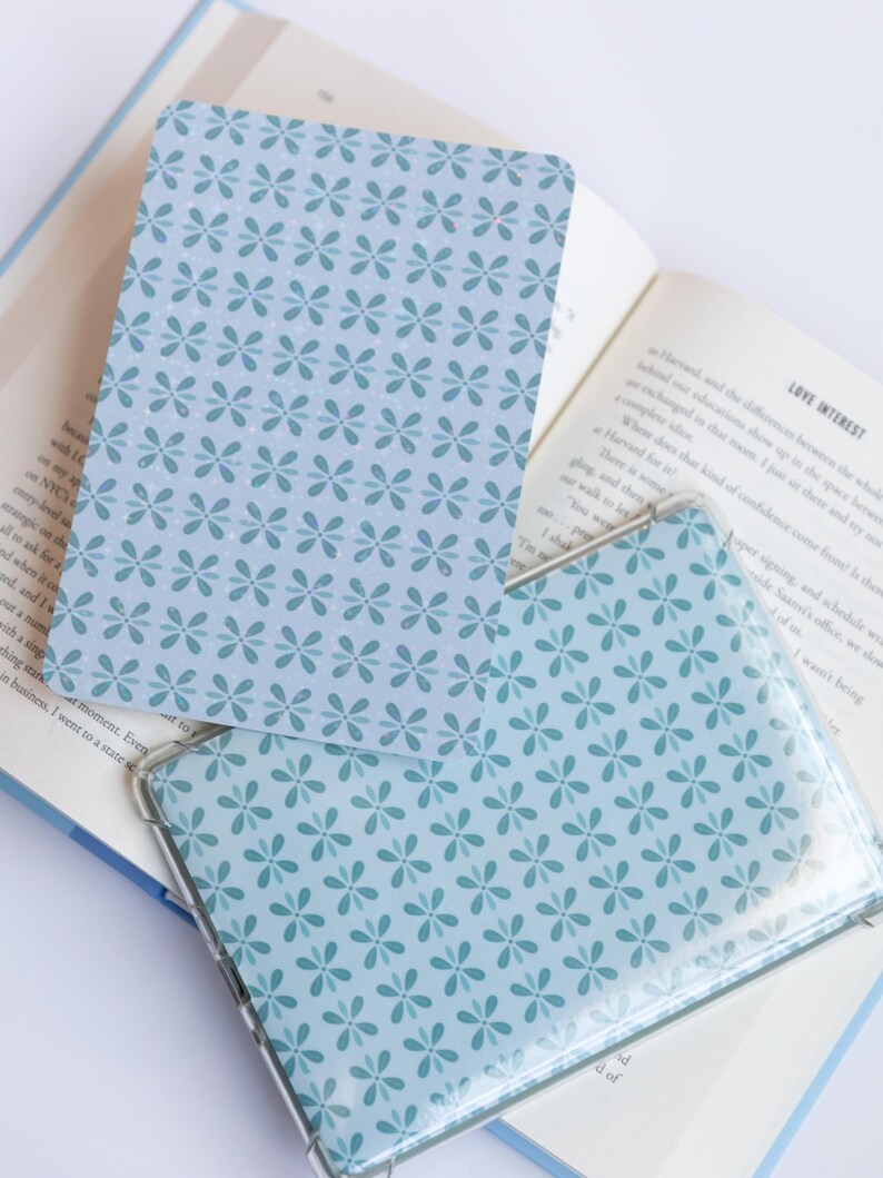 May include: Two light blue floral patterned cards, one inside a clear plastic sleeve, rest on an open book. The cards and sleeve feature a repeating pattern of small, stylized flowers in a darker shade of blue.