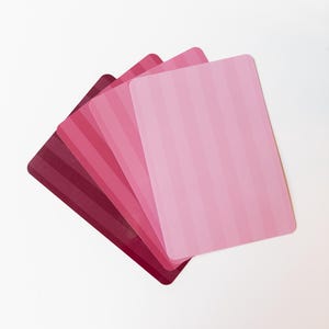 May include: A collection of five rectangular cards in varying shades of pink, arranged in a fanned-out pattern. Each card features vertical stripes, with colors ranging from deep burgundy to light pink.