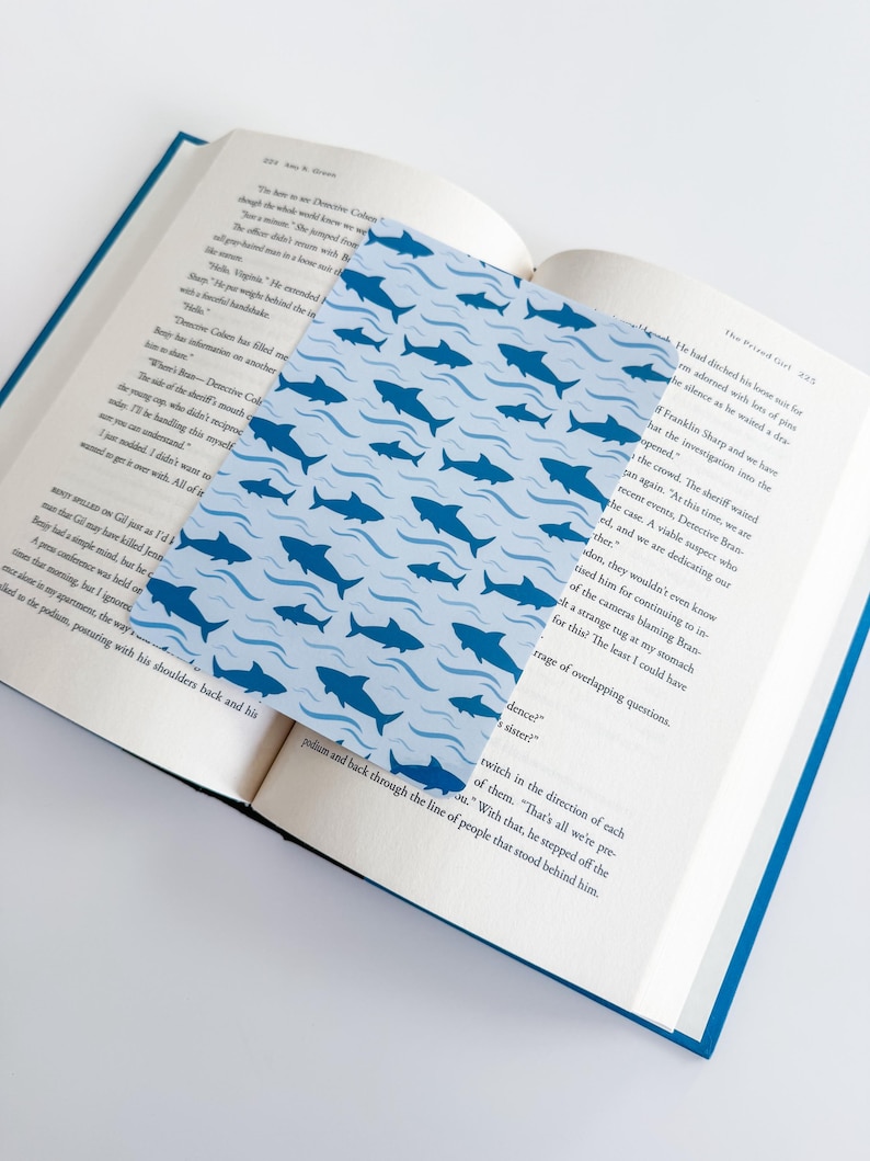 K&ouml;nnte beinhalten: Ein rechteckiges Lesezeichen mit einem sich wiederholenden Muster aus dunkelblauen Haien, die zwischen hellblauen Wellenlinien schwimmen. Das Lesezeichen liegt auf den offenen Seiten eines Buches mit gedrucktem Text, Seite 224 und 225.