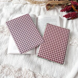 May include: Two rectangular e-reader or tablet covers featuring a dark maroon and white gingham check pattern. One cover is placed flat on an open book, while the other is positioned to the right, showing a clear protective frame around the edges.