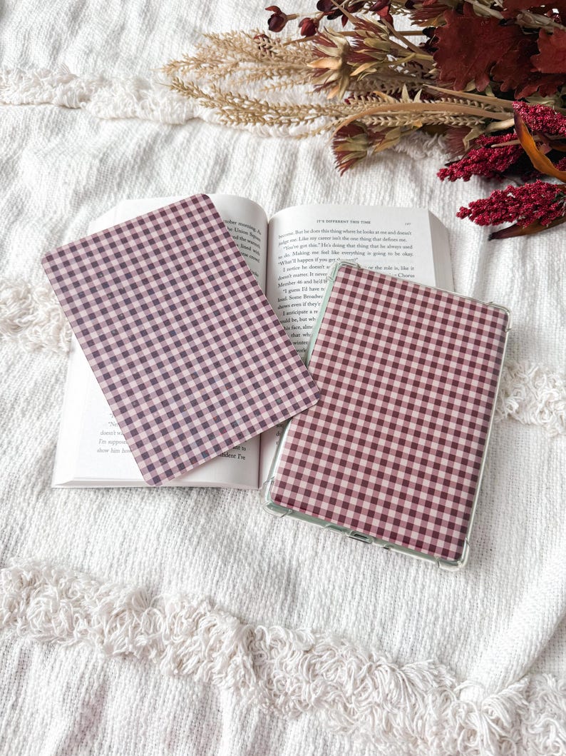 May include: Two rectangular e-reader or tablet covers featuring a dark maroon and white gingham check pattern. One cover is placed flat on an open book, while the other is positioned to the right, showing a clear protective frame around the edges.
