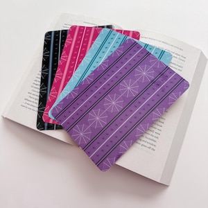 May include: Four rectangular bookmarks with rounded corners, each featuring a vertical striped pattern with white spiderwebs and small star icons. The bookmarks are colored in black, magenta, light blue, and purple, layered over an open book.