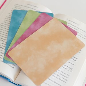 May include: A stack of colorful, textured bookmarks in shades of blue, green, pink, and peach, fanned out over an open book. The bookmarks have a watercolor-like effect, adding a soft, artistic touch to the reading experience.