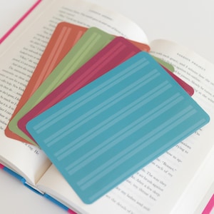 May include: A collection of colorful, striped bookmarks fanned out over an open book. The bookmarks are in shades of orange, green, red, and blue. The book has white pages and a pink cover.
