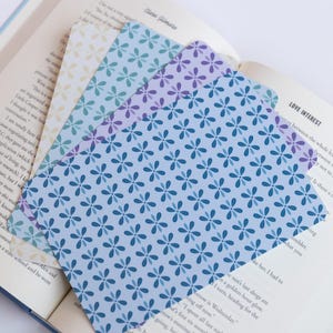 May include: Set of three floral patterned bookmarks in shades of blue, purple, and cream, resting on the open pages of a book. The bookmarks feature a repeating flower design. The book has text on the pages.