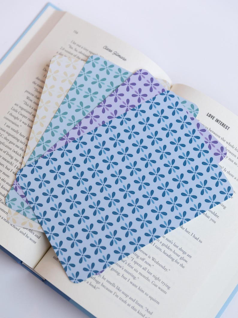 May include: Three floral patterned bookmarks in shades of blue, purple, and cream, on the open pages of a book. The bookmarks have a repeating flower design. The book has text on the pages.