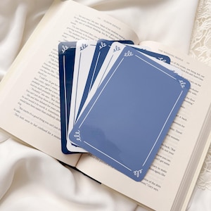 May include: A stack of blue and white cards with decorative borders, resting on an open book. The cards have a light blue background with white borders and the word "ele" in the corners. The book's pages are visible.
