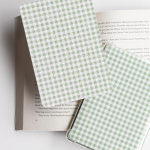 May include: A green and white gingham patterned phone case and a matching card are displayed open on a book. The phone case is clear with a green and white checkered design. The card has the same pattern.