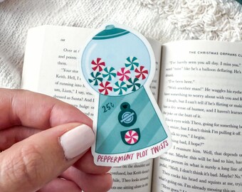 May include: A sticker featuring a teal gumball machine design with red and white peppermint candies. The sticker has the text "25¢" and "Peppermint Plot Twists" on it. The sticker is placed on an open book.