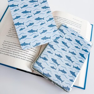 K&ouml;nnte beinhalten: Eine rechteckige E-Reader-H&uuml;lle und ein passendes flaches Cover mit einem sich wiederholenden Muster aus dunkelblauen Haien, die durch hellblaue Wellenlinien schwimmen. Die Gegenst&auml;nde liegen auf einem aufgeschlagenen Buch mit sichtbarem Text.