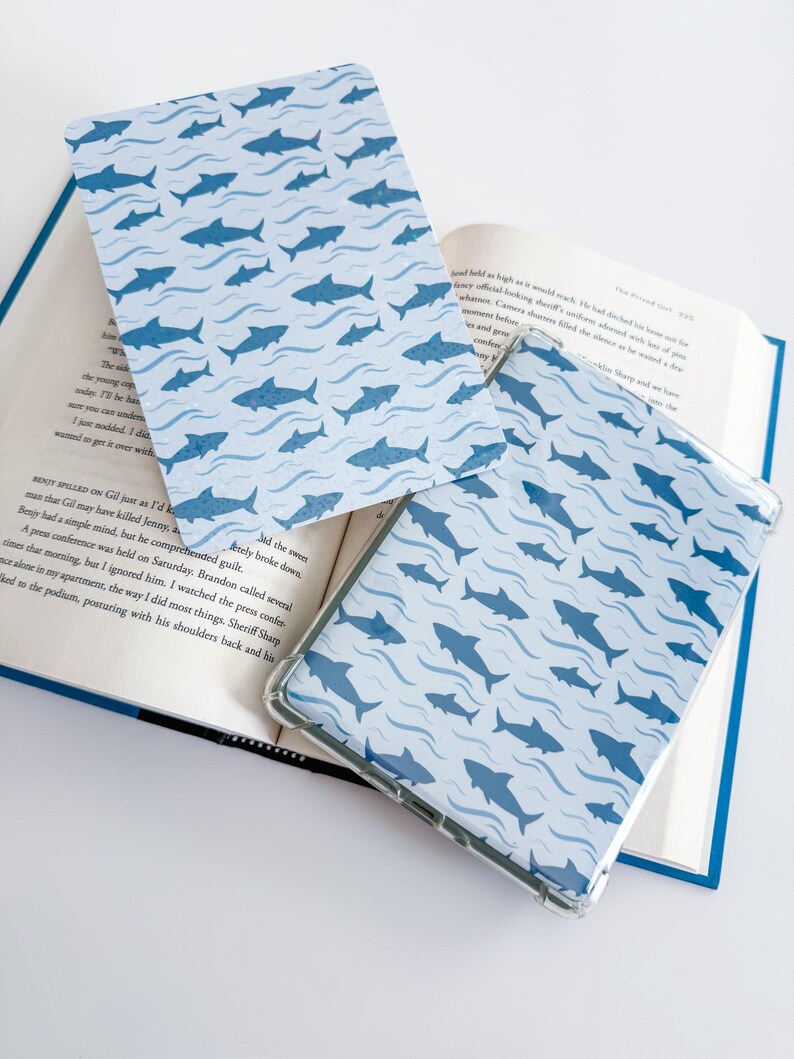 K&ouml;nnte beinhalten: Eine rechteckige E-Reader-H&uuml;lle und ein passendes flaches Cover mit einem sich wiederholenden Muster aus dunkelblauen Haien, die durch hellblaue Wellenlinien schwimmen. Die Gegenst&auml;nde liegen auf einem aufgeschlagenen Buch mit sichtbarem Text.