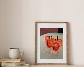 Könnte beinhalten: Ein gerahmter Druck mit sechs Aperol Spritz Cocktails mit schwarzen Strohhalmen auf einem hellgrünen Tisch. Die Getränke befinden sich in Stielgläsern, gefüllt mit orangener Flüssigkeit und mit Orangenscheiben garniert. Der Hintergrund zeigt eine Backsteinterrasse.