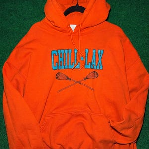 May include: Orange hoodie with the text "CHILL LAX" in blue and crossed lacrosse sticks. The hoodie has a drawstring hood and a front pocket. The garment is hanging on a black hanger against a green background.