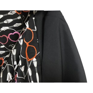 Puede incluir: Camisa de manga larga negra y blanca con un dise&ntilde;o bordado en blanco y negro de caras con gafas y flechas. La camisa tiene una tela negra en el lado derecho.