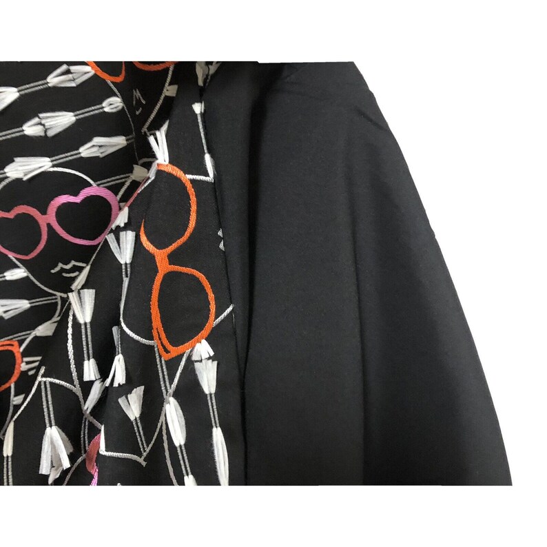 Puede incluir: Camisa de manga larga negra y blanca con un dise&ntilde;o bordado en blanco y negro de caras con gafas y flechas. La camisa tiene una tela negra en el lado derecho.
