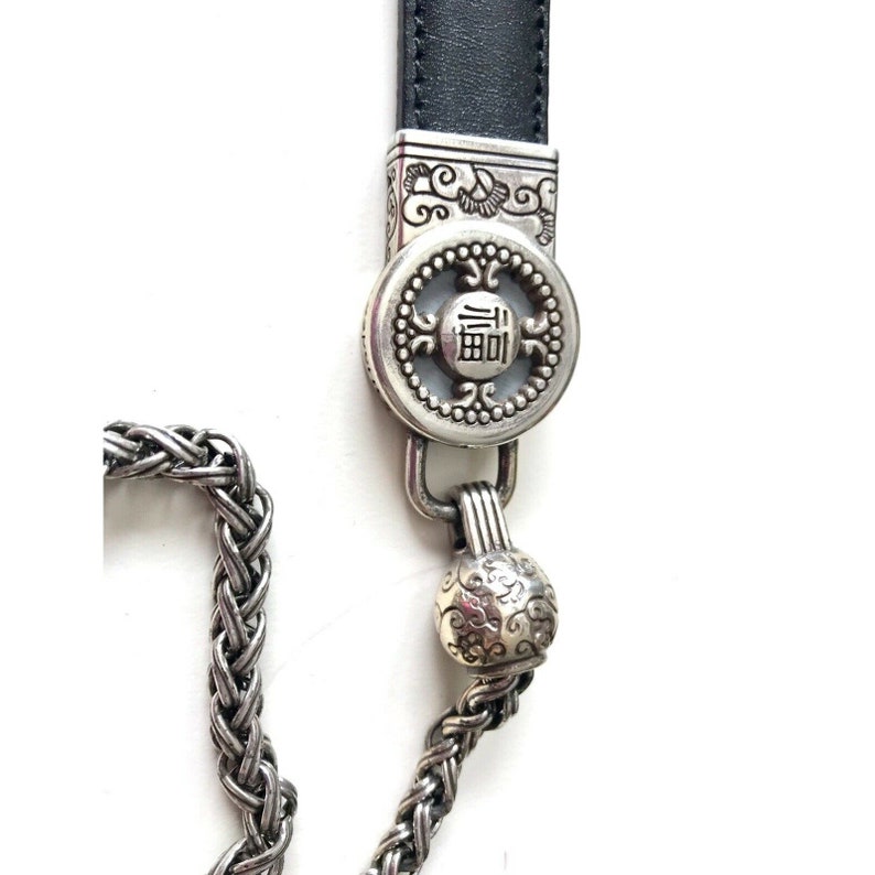 K&ouml;nnte beinhalten: Eine silberne Kette mit einem dekorativen Verschluss, der das chinesische Schriftzeichen f&uuml;r "Gl&uuml;ck" zeigt, und einem kleinen, verzierten Kugelanh&auml;nger. Die Kette ist an einem schwarzen Lederband befestigt.