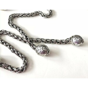 K&ouml;nnte beinhalten: Eine silberne Kettenkette mit zwei dekorativen Endkappen. Die Kette ist geflochten und die Endkappen haben ein florales Design.