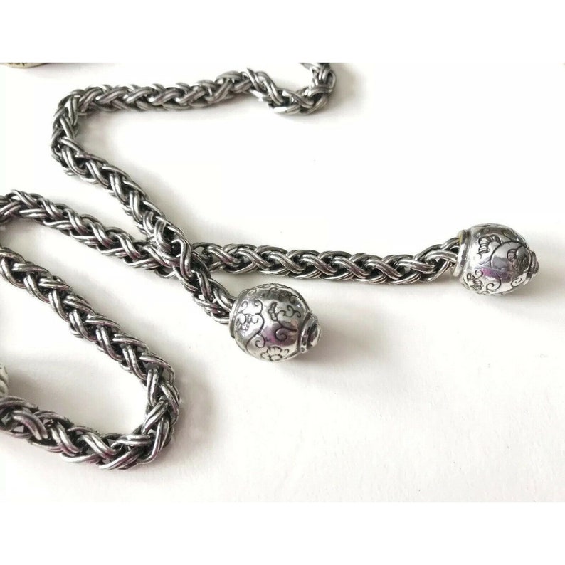 K&ouml;nnte beinhalten: Eine silberne Kettenkette mit zwei dekorativen Endkappen. Die Kette ist geflochten und die Endkappen haben ein florales Design.