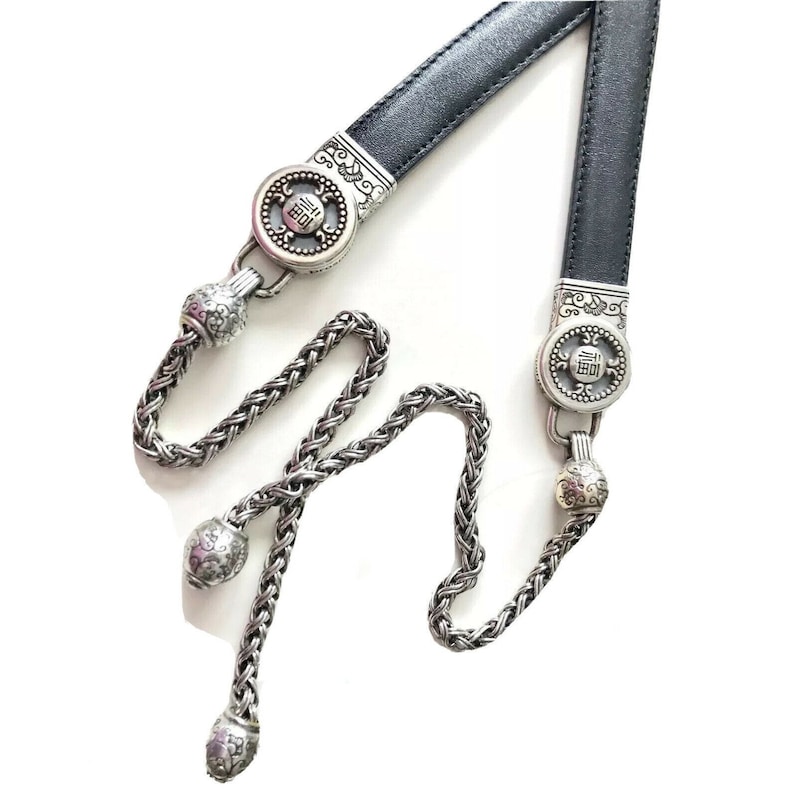 K&ouml;nnte beinhalten: Ein schwarzer Lederg&uuml;rtel mit silbernen Kettenquasten. Der G&uuml;rtel hat zwei silberne Schnallen mit verzierten Designs und das chinesische Schriftzeichen f&uuml;r "Gl&uuml;ck" ist auf jeder Schnalle eingraviert.