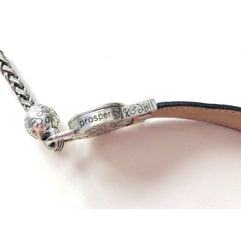 K&ouml;nnte beinhalten: Ein silbernes Armband mit einem schwarzen Lederband und einem silbernen Anh&auml;nger, auf dem "prosperity &" steht.