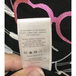 Puede incluir: Etiqueta blanca con texto negro sobre un fondo negro con un dise&ntilde;o de coraz&oacute;n rosa y gafas. La etiqueta dice "DO NOT WASH, DO NOT BLEACH. DO NOT TUMBLE DRY, LOW IRON DRY CLEAN ONLY NE PAS LAVER A LA MACHINE NE PAS JAVELLISER NE PAS SECHER A LA MACHINE REPASSER AL FER FROID NETTOYER A SEC SEULEMENT FOR BU IMPORTED BY ANTHROPOLOGIE 24 MARKET PLACE SHOE FLOOR LONDON UK W1B4DG STYLE # 168"