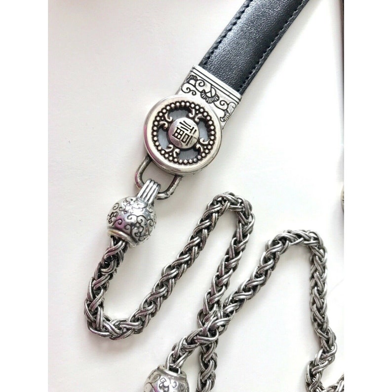 K&ouml;nnte beinhalten: Ein silberner Ketteng&uuml;rtel mit einem schwarzen Lederriemen und einem runden Anh&auml;nger mit chinesischen Schriftzeichen. Der Anh&auml;nger hat einen dekorativen Rand und ein erhabenes Design.