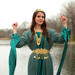 Könnte beinhalten: Eine Frau in einem tealgrünen Kleid und einer passenden Jacke mit langen Ärmeln, verziert mit einer goldfarbenen Krone, Halskette und Gürtel. Das Kleid hat ein langes, fließendes Design mit einem goldfarbenen Gürtel mit baumelnden Münzen. Die Jacke hat lange Ärmel mit tealfarbenen Bändern.