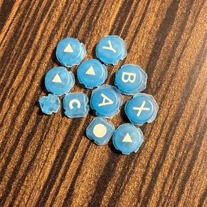 May include: A collection of light blue game controller buttons with white symbols and letters, including A, B, X, Y, C, and a white triangle. The buttons are scattered on a brown wooden surface.