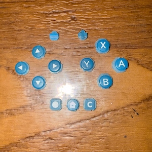 May include: A collection of light blue game console buttons, including directional arrows, lettered buttons (A, B, C, X, Y), and other function buttons. The buttons are scattered on a wooden surface.