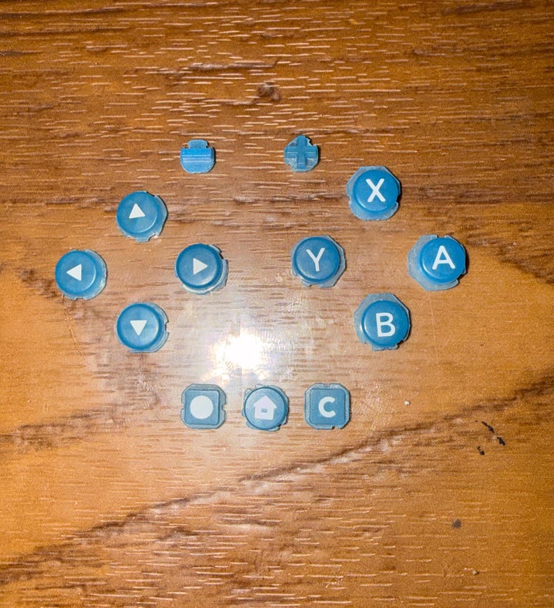 May include: A collection of light blue game console buttons, including directional arrows, lettered buttons (A, B, C, X, Y), and other function buttons. The buttons are scattered on a wooden surface.