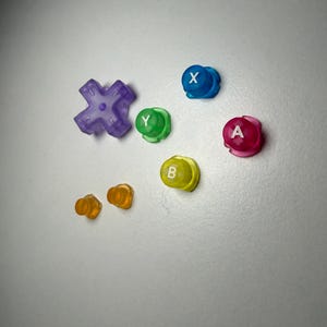May include: A selection of brightly coloured video game controller buttons and thumbsticks. The buttons are in various colours: purple, blue, green, yellow, and pink, each with a white letter. Two orange thumbsticks are also present.