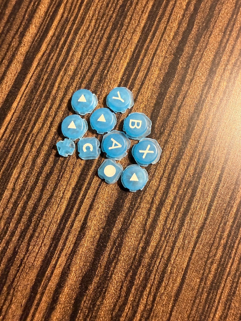 May include: A collection of light blue game controller buttons with white symbols and letters, including A, B, X, Y, C, and a white triangle. The buttons are scattered on a brown wooden surface.