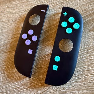 以下が含まれることがあります： 2つの黒いゲームコントローラー。片方は紫色のボタンに白い記号、もう片方はターコイズ色のボタンに白い記号があります。ゲーム機用のコントローラーです。