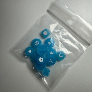 May include: A clear plastic bag filled with several small, translucent blue keycaps. Each keycap has a white symbol or letter, including the letters "B" and "C", and directional arrows. The keycaps are scattered inside the bag.