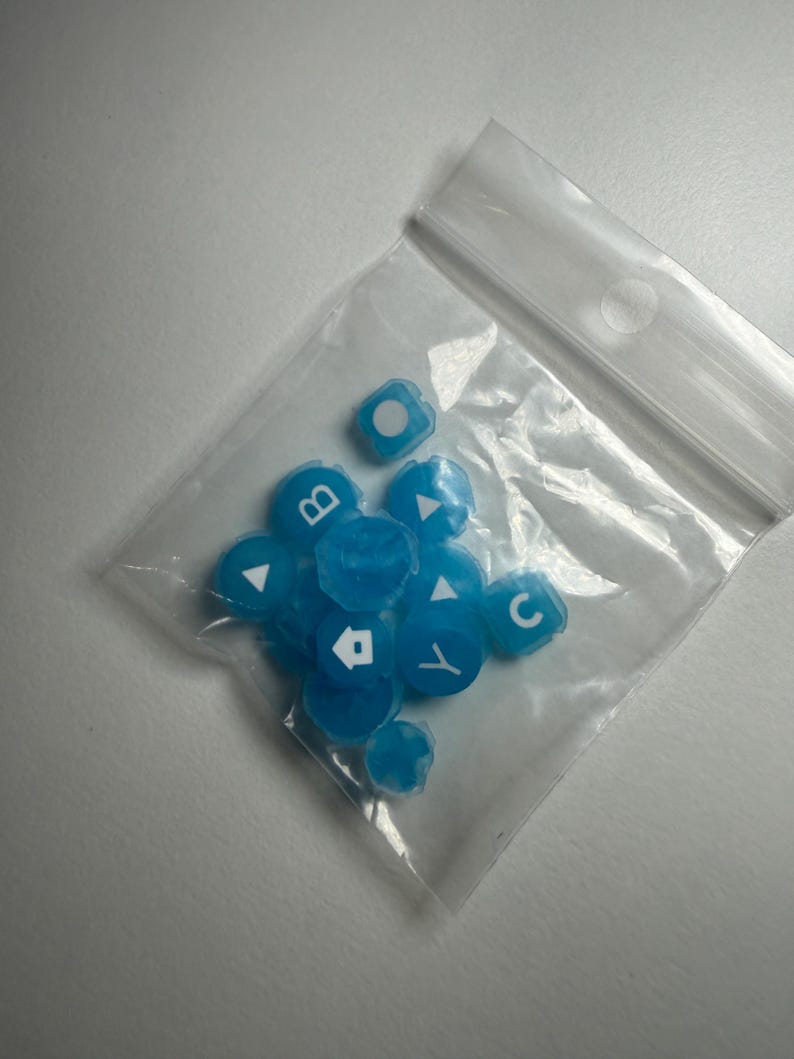 May include: A clear plastic bag filled with several small, translucent blue keycaps. Each keycap has a white symbol or letter, including the letters "B" and "C", and directional arrows. The keycaps are scattered inside the bag.