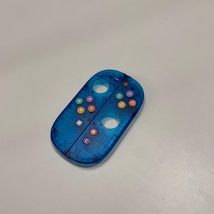 May include: A transparent blue video game controller with colored buttons and directional controls. The controller has a series of colored buttons, including red, yellow, green, and purple. The controller is designed for a gaming console.