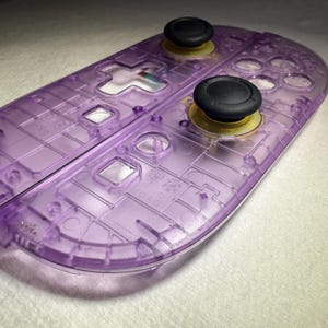 May include: A transparent purple game controller shell with black joysticks. The controller has various button and port cutouts, and a split design. The shell is made of a clear, translucent plastic.