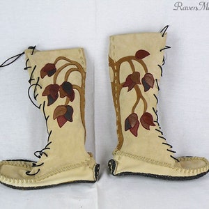 Op de afbeelding: Een paar kniehoge, crèmekleurige mocassinlaarzen. De laarzen hebben een bloemenmotief in bruin, bordeauxrood en beige. Zwarte veters lopen over de voorkant. De zolen zijn donker en de laarzen zijn gemaakt van zacht materiaal. De tekst "RavenMountain.ca" staat rechtsboven.