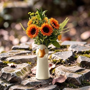 Op de afbeelding: Een witte keramische vaas in de vorm van een ganzenkop, met een geeloranje snavel en zwarte ogen, gevuld met een levendig boeket van oranje zonnebloemen, kleine groene knopbloemen en diverse groene bladstelen.