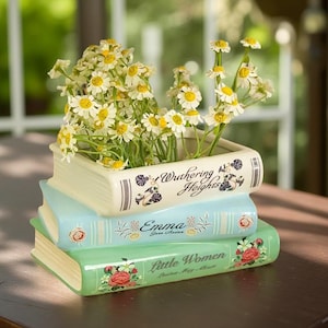 Könnte beinhalten: Dekorativer Keramik-Blumentopf in Form eines Bücherstapels mit den Titeln "Wuthering Heights", "Emma" und "Little Women". Der Topf ist mit kleinen weißen und gelben Blumen gefüllt. Die Bücher sind in Creme, Hellblau und Grün gehalten.