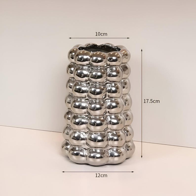 May include: A silver vase with a bubbly texture. The vase is 17.5 cm tall, 12 cm wide at the base, and 10 cm wide at the top. The vase has a reflective, metallic finish and a unique, modern design. This decorative vase is suitable for home decor.